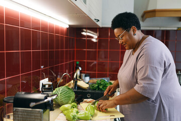 Side view of happy plus size african american senior woman cooking healthy delicious veggie stew...