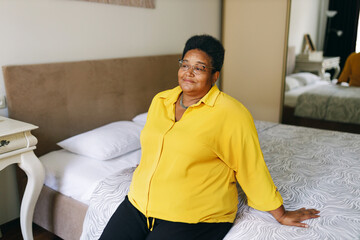 Big plus size african american grandmother in yellow clothes and eyeglasses sitting on bed, leaning on hands with thoughtful positive facial expression, recollecting best memories of her life