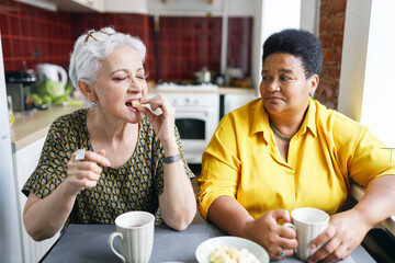 Fototapeta na wymiar Senior caucasian female trying sweats and enjoying delicious taste with closed eyes sitting at kitchen table next to her african american woman friend on diet, drinking coffee or tea together