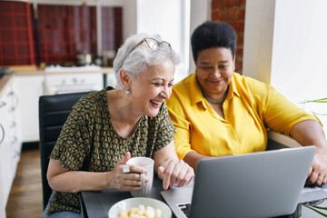 Image of two senior senior female besties of diverse ethnicity watching funny show or comic series while drinking coffee at kitchen table, caucasian woman laughing out loud looking at screen