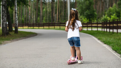 Seasonal children's sports. Healthy childhood lifestyle. Summer holidays. Little girl, child riding a scooter on the road in the park outdoors.