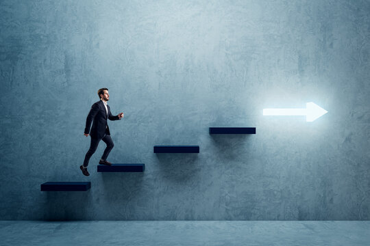 Side View Of Young Business Man Climbing Illuminated Arrow Stairs To Success On Concrete Wall Background In Interior. Financial Growth, Career Development And Forward Concept.