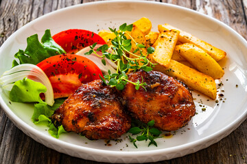 Roasted chicken thighs with French fries and fresh vegetables on wooden table
