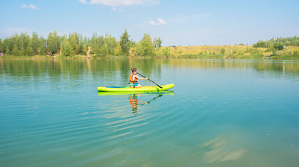 a boy rides a light green SUP board along a mountain river on summer vacation. A 10-year-old child goes in for water sports, in a life jacket, stands on a SUP surfboard.