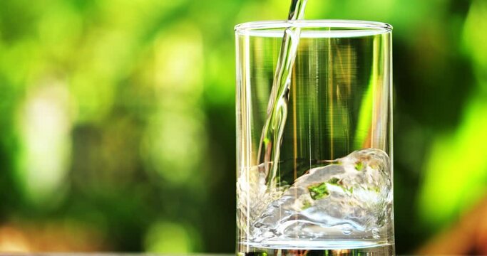 Pouring water concept. Close-up person pouring the drinking water into transparent glass in slow motion. Nature background, freshness and healthy.