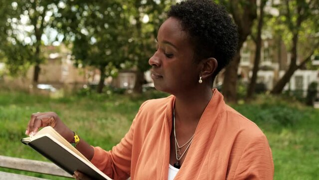 Black woman reading a book on a bench park.