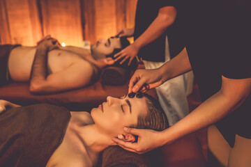 Caucasian couple enjoying relaxing anti-stress head massage with hot stone and pampering facial beauty skin recreation leisure in warm candle light ambient salon spa in resort or hotel. Quiescent