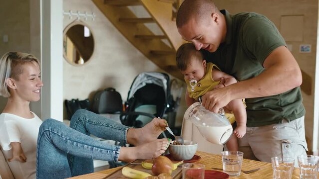 Happy young family with a litle baby is preparing breakfast at the table. A woman living with Phocomelia syndrome holds a spoon with her foot while her husband pours milk while preparing breakfast t