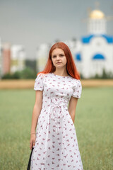 Obraz premium Portrait of a young beautiful red-haired girl in a dress in a field on the background of the church.