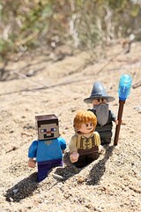 Obraz premium LEGO Minecraft figure of Steve walking on sandy beach with LEGO Lord Of The Rings wizard Gandalf and hobit Samwise Gamgee figures, summer daylight sunshine. Some arid foliage and beach in background. 