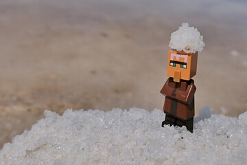 Obraz premium LEGO Minecraft figure of Villager mob standing on pile of salt next to salt manufacturing field with piece of salt on his head, sunlit by summer daylight sunshine. 