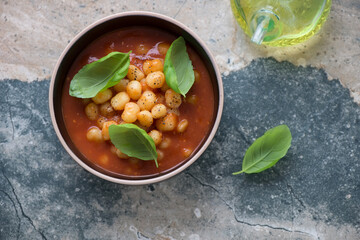 Tomato gnocchi soup served in a bowl with fresh green basil, above view on a grey and beige stone background, horizontal shot
