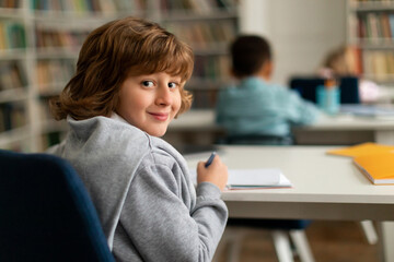 Smart male elementary school pupil, sitting at desk in classroom, turning back and smiling at camera during lesson
