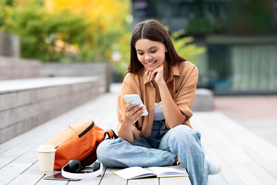 Happy young woman university student using smartphone app sitting outdoors