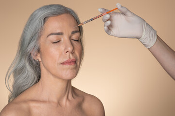 Skin lifting concept. Headshot of senior woman receiving beauty filler injection in forehead, beige background