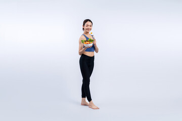 Young sporty Asian woman in sportswear holding salad bowl fill with vibrant of fruit and vegetable. Natural youthful and fit body lifestyle with balance nutrition on isolated background. Vigorous