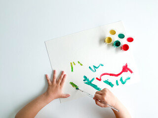 small child draws with paints and brush on white table.