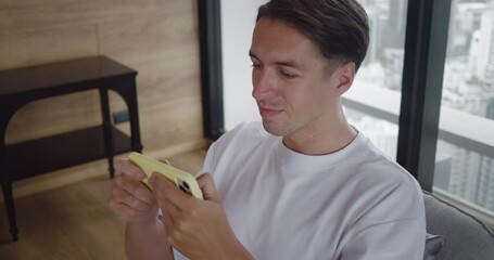 30s man in white shirt plays mobile game on a smartphone, sitting at home in a chair against the backdrop of a window overlooking big city. Male gamer enjoying mobile app game racing simulator at home