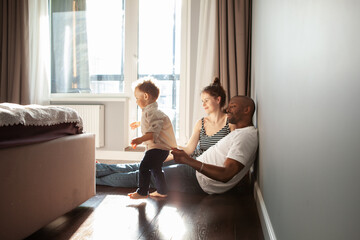 Diverse multiracial family playing with child son at home, authentic family life, unposed, spontaneous moments. Baby learning walk and dad supports him, helps
