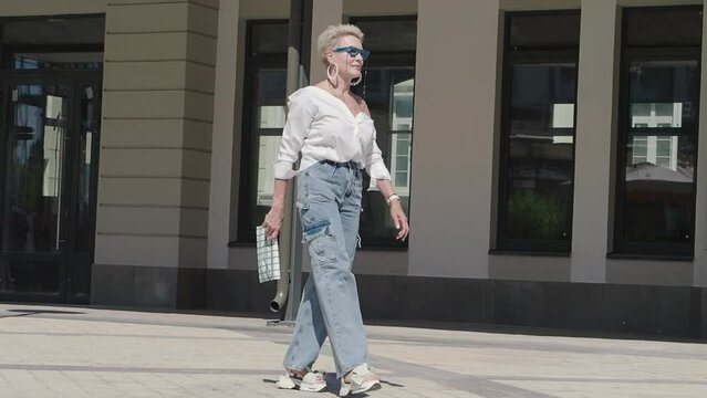 Beautiful mature woman walks outdoors in city street dressed trendy white shirt, jeans cargo pants, Thick platform sandals with open toe, clutch. Urban street style fashion