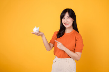 Smiling Asian woman in her 30s holding piggy bank, pointing to it, on vibrant yellow background. Wealth and saving concept.