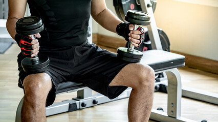 Athlete man in black sportswear wearing sport gloves sitting on workout bench at weightlifting station holding dumbbells prepare for lifting in fitness gym. Weight training and bodybuilding concepts - obrazy, fototapety, plakaty