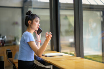 Asian young woman using mobile phone to call someone at coffee shop. Looking outside the window.