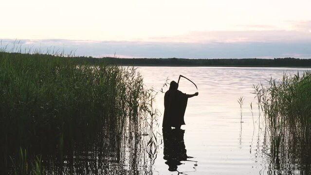 Death tests the scythe while standing in a pond before looking for a new victim at dusk. Silhouette of an unrecognizable person.