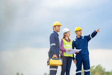 A team of 3 male and female engineers has a blueprint for a project meeting. Look and point your finger at industrial areas. Wear a safety helmet. Mechanic's vest and uniform with toolbox