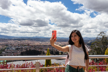 mujer latina de cabello largo negro y blusa blanca tomandose una selfie en un mirador en la ciudad...