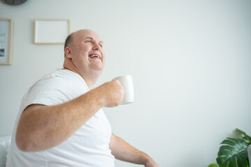 Comfortable Awakening: Chubby Caucasian Man Enjoys Morning Coffee on Bed in Cozy Home Environment