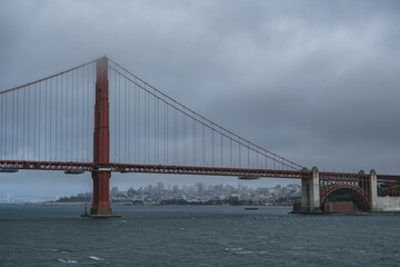 Fototapeta na wymiar Red metal steel suspension bridge Golden Gate Bridge in San Francisco Bay Area with coast and city downtown skyline silhouette on cloudy day from outdoor cruiseship cruise ship deck