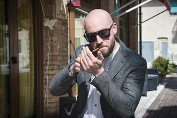 Young man with a shaved head, beard, and sunglasses, lights up a cigar.