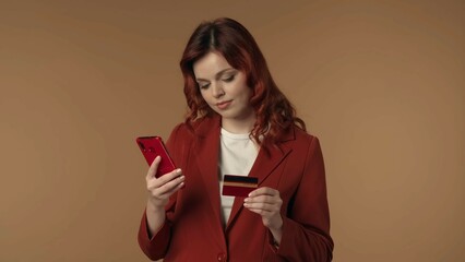 Naklejka premium Medium isolated shot of a joyful, satisfied and relaxed young woman looking at her phone and holding a credit card, completing an online purchase.
