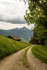 Road Leading By a Building Sitting Along a Hill of the Swiss Alps in Switzerland in the Summer with Mountains Peaking Through Clouds in the Background