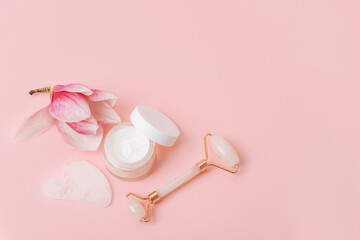 Rose quartz crystal facial roller and gua sha scraper, face cream, magnolia flower on pink background. Facial massage kit for lifting therapy. Top view, flat lay, copy space