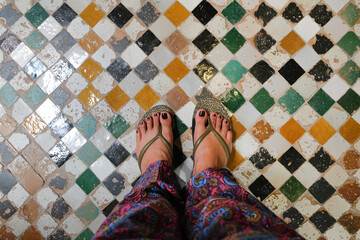 Female feet wearing slippers and printed bohemian pants stands on old, colorful checkered tiles. Floor inside a traditional riad in Fez, Morocco. Horizontal travel background and copy space.