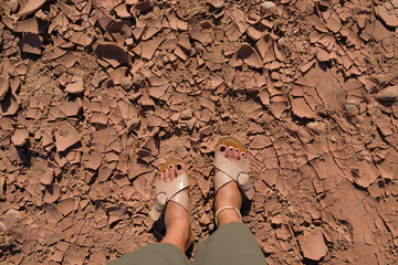 Female feet with sandals stands on cracked, parched soil in Ait Ben Haddou, a historic clay village in Morocco. Global warming concept. Person steps on dry earth. Dehydrated ground. Travel background.