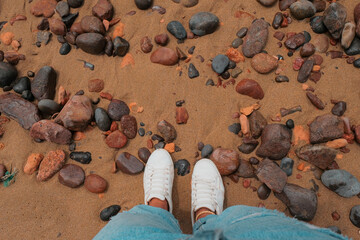 Looking down at sand and rocks in Legzira Beach. Rugged coastline in the Tiznit Province of Morocco, Africa. Perspective shot. Ripped blue jeans and white sneakers. Travel background. Summer day.