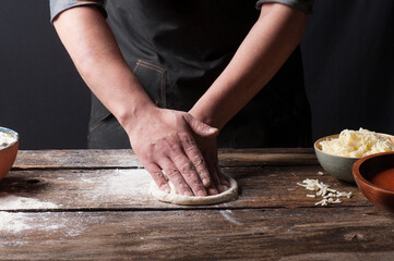 Dark and moody photo of a man hand rolling pizza dough on a vintage wooden counter with bowls of flour, tomato sauce and cheese