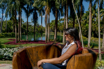 Mulher sentada em grande tronco de árvore, trabalhado rusticamente e feito de banco, linda paisagem ao fundo com lago, muitas palmeiras e vegetação, no museu a céu aberto de Minas Gerais - 85