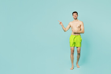 Full body young sexy man wears green shorts swimsuit relax near hotel pool point index finger aside on area mock up isolated on plain light blue background. Summer vacation sea rest sun tan concept.