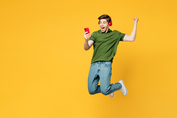 Fototapeta na wymiar Full body young excited happy man wear green t-shirt casual clothes jump high listen to music in headphones use mobile cell phone do winner gesture isolated on plain yellow background studio portrait