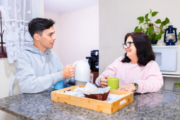Mature woman and adult son enjoy a traditional Brazilian coffee break at a table, sharing lively conversation in a typical Brazilian home