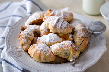 A plate with small bagels, croissants sprinkled with powdered sugar, a kitchen towel in the frame, a bottle of milk. Homemade baking.