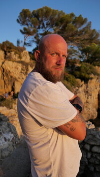 Striking portrait of a man with blue eyes and a beard, set against the picturesque landscape of Cap d'Antibes, France. His tattoos tell a story, embodying a blend of rugged charm and natural beauty