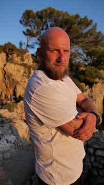 Striking portrait of a man with blue eyes and a beard, set against the picturesque landscape of Cap d'Antibes, France. His tattoos tell a story, embodying a blend of rugged charm and natural beauty