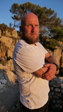 Striking portrait of a man with blue eyes and a beard, set against the picturesque landscape of Cap d'Antibes, France. His tattoos tell a story, embodying a blend of rugged charm and natural beauty