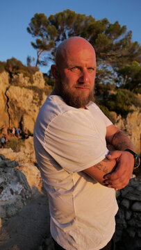 Striking portrait of a man with blue eyes and a beard, set against the picturesque landscape of Cap d'Antibes, France. His tattoos tell a story, embodying a blend of rugged charm and natural beauty