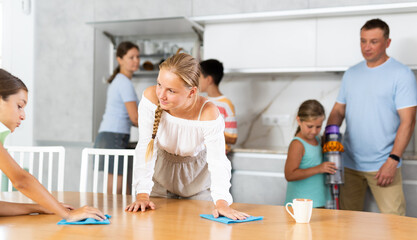 Two teenage girls wipe table and help their mother clean up in kitchen, wife with husband and...